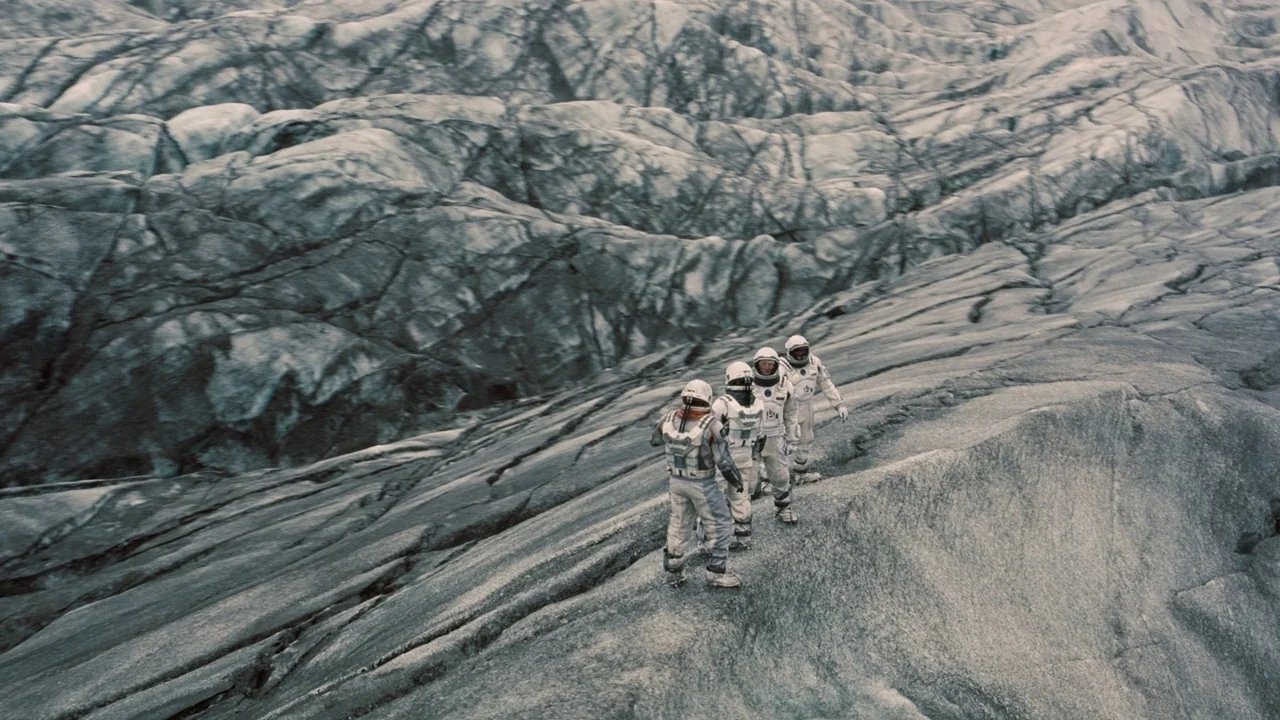 Interstellar (2014) movie still: wide — A vast, rugged expanse of grey ice forms a desolate, ridged landscape. Four astronauts in white pressurized…