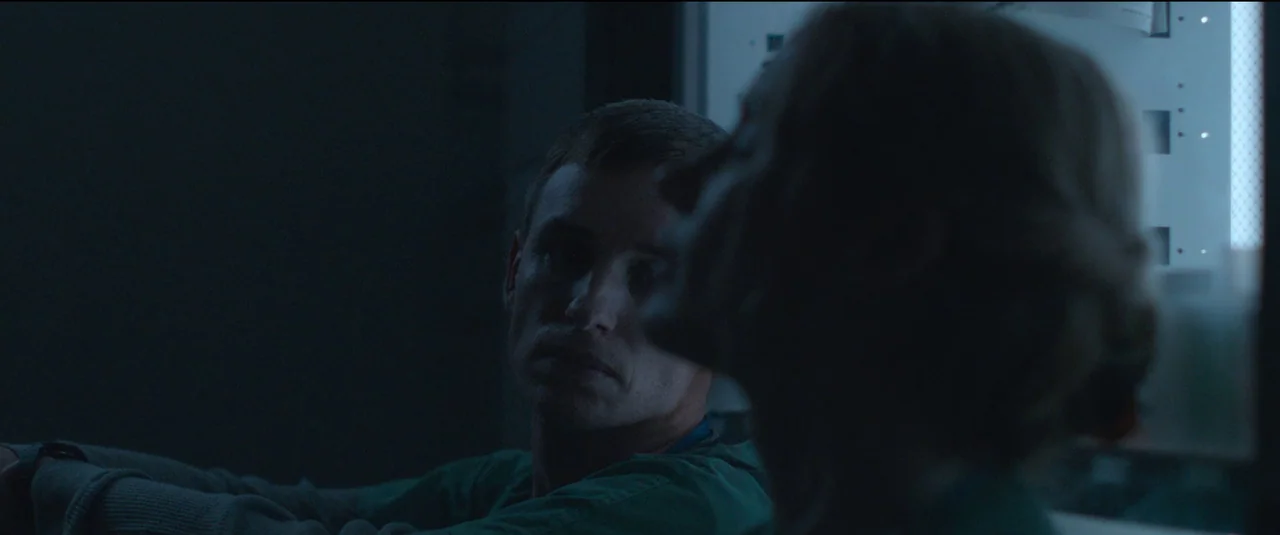 The Good Nurse (2022) movie still: medium — A dimly lit room contains medical equipment with blue-tinted screen displays. A man in green scrubs…