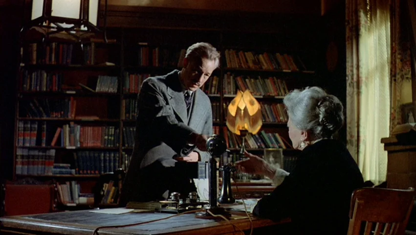 Hammett (1982) movie still: medium — A dimly lit home office features floor-to-ceiling bookshelves packed with books and a desk. A man in a suit…