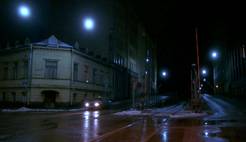 Night on Earth (1991) movie still: wide — A dark city street corner at night features a pale yellow corner building. Tram tracks cross a wet asphalt…