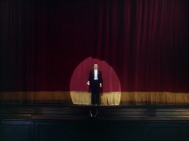 The Red Shoes (1948) movie still: fullbody — A dark empty theater stage features heavy floor-to-ceiling red velvet curtains with gold trim. A single…