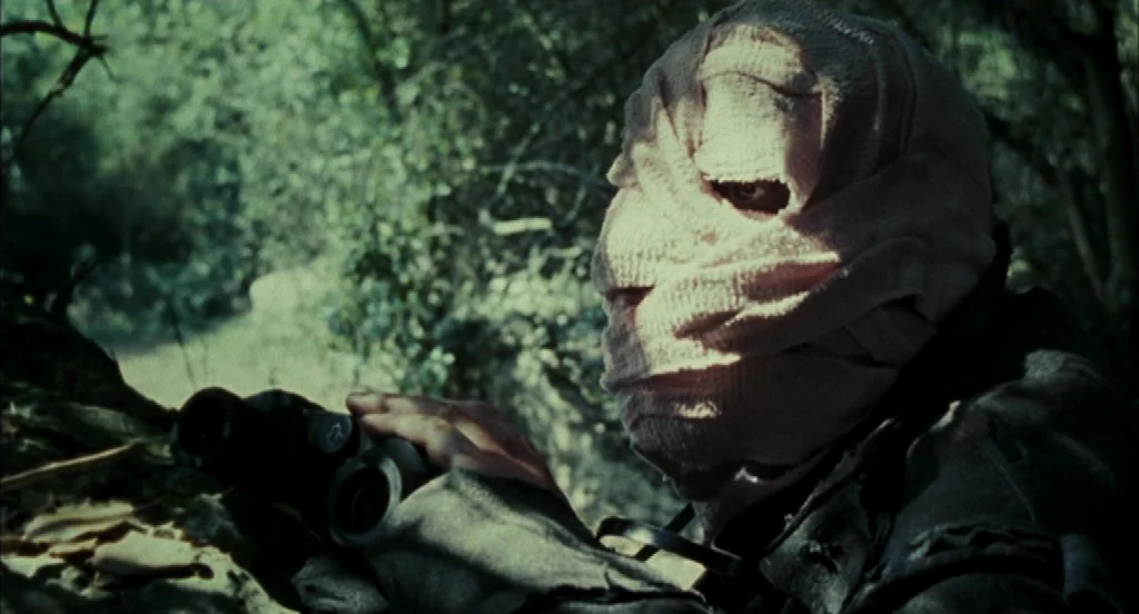 Timecrimes (2007) movie still: closeup — A person with their head completely wrapped in medical bandages hides within dense woodland. They peer…