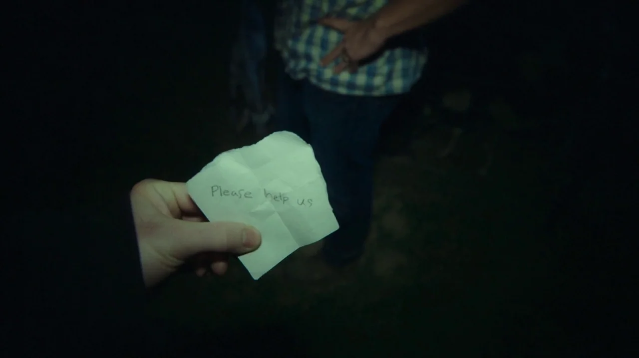The Sacrament (2013) movie still: closeup — A dark outdoor setting where a person presents a handwritten note in the foreground. A second figure…