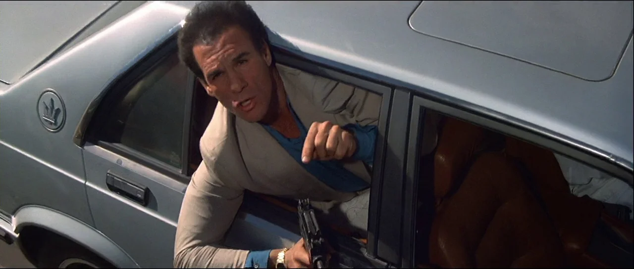 Licence to Kill (1989) movie still: medium — A silver vehicle is traveling on a road during the daytime. The driver leans out the open passenger…