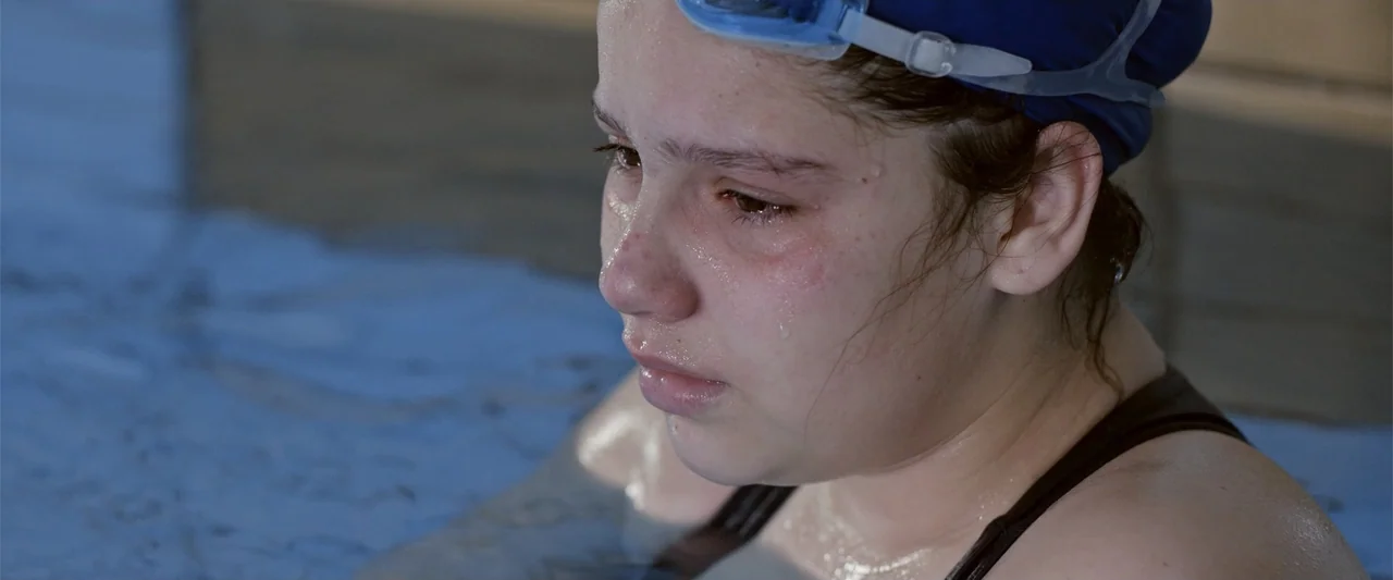 Dirty Little Secret (1998) movie still: closeup — A teenage girl stands waist-deep in the blue water of an indoor pool. She wears a black swimsuit…