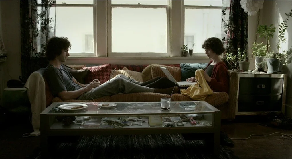 The Future (2011) movie still: medium — A furnished interior space with a tan couch placed in front of three large windows. Two individuals sit…