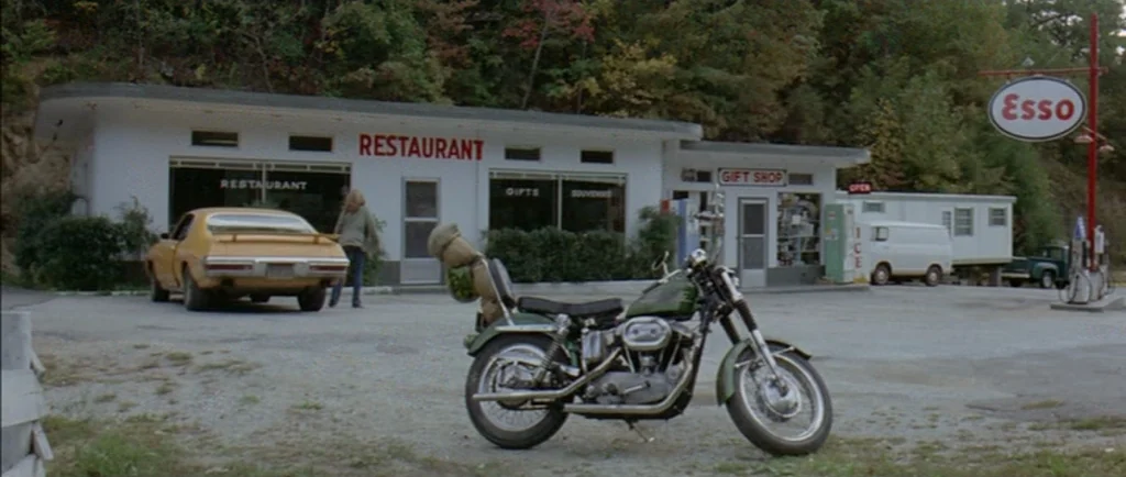 Two Lane Blacktop (1971) movie still: wide — A rural roadside building complex features a restaurant, a gift shop, and an Esso sign. A woman walks…