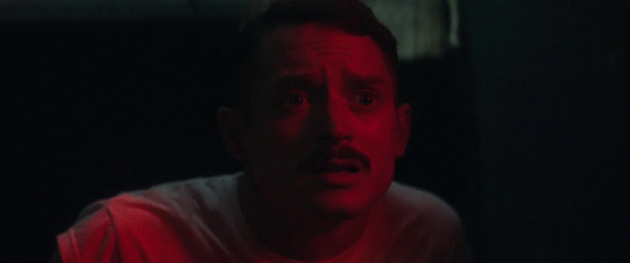 Come To Daddy (2019) movie still: closeup — A man stands in a dimly lit, dark space bathed in harsh crimson light. He looks intently towards the…