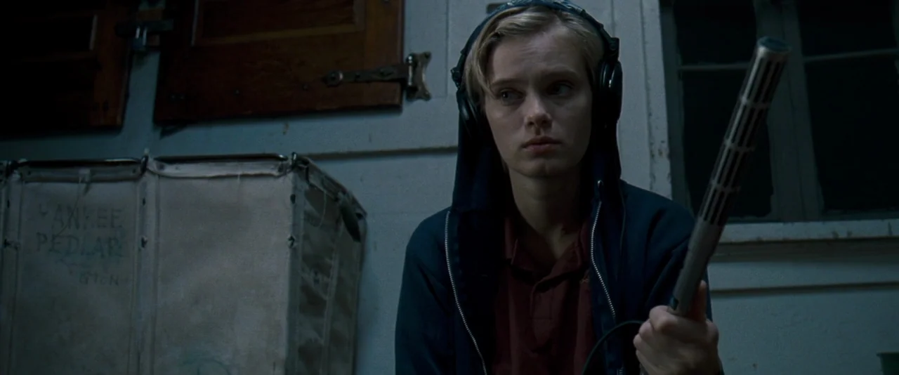 The Innkeepers (2011) movie still: medium — An interior space with a large canvas laundry cart and a window with slats. A young woman wears…