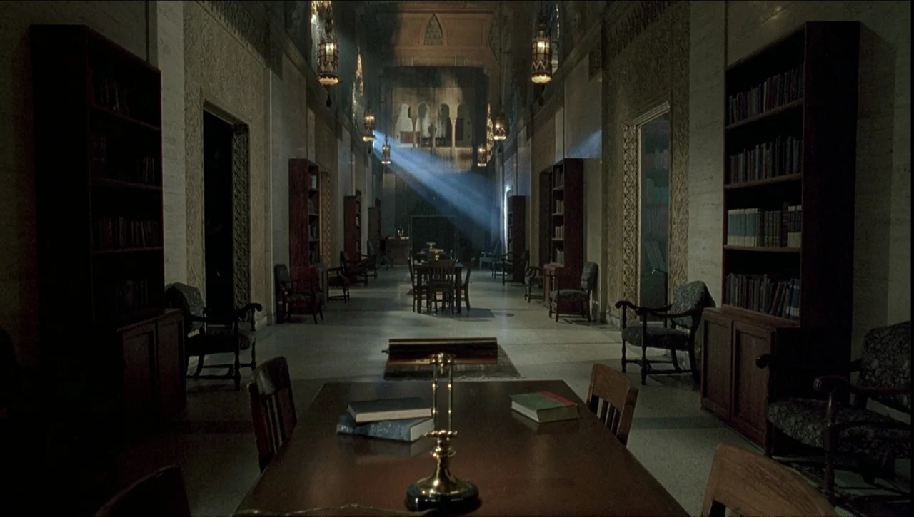 Necronomicon: Book of Dead (1993) movie still: establishing — A long, dimly lit institutional hallway features wooden bookcases and chairs lining…