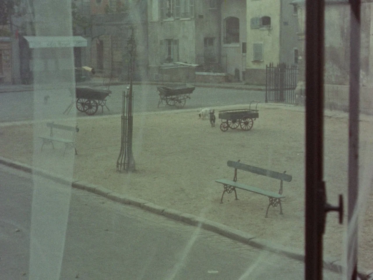 Mon Oncle (1958) movie still: establishing — An empty cobblestone square features iron benches, wooden carts, and a metal tree cage. Two small dogs…