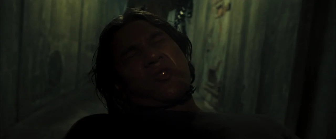 The Unholy (2022) movie still: closeup — A narrow, dark interior corridor with textured, aged walls features a man centered in frame. The man leans…