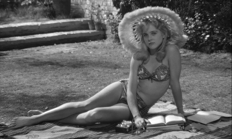 Lolita (1962) movie still: fullbody — A woman sits on a patterned blanket spread across a manicured lawn. She poses while wearing a bikini and a…