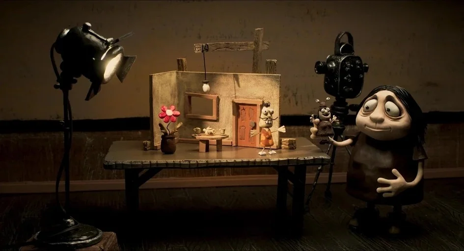 Memoir of a Snail (2024) movie still: medium — A stop-motion puppet stands on a surface beside a miniature set and camera. The character holds a…