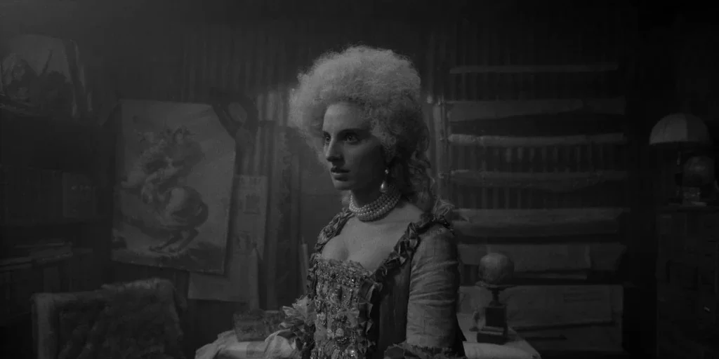 El Conde (2023) movie still: medium — A dark study contains a woman wearing an 18th-century costume and a powdered wig. She gazes off-camera while…