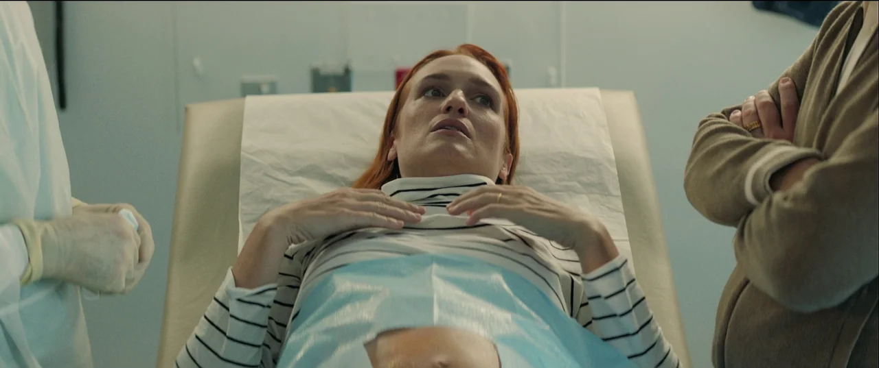 Birth/Rebirth (2023) movie still: medium — A clinical exam room features pale walls and a paper-covered medical table. A woman lies on the table…