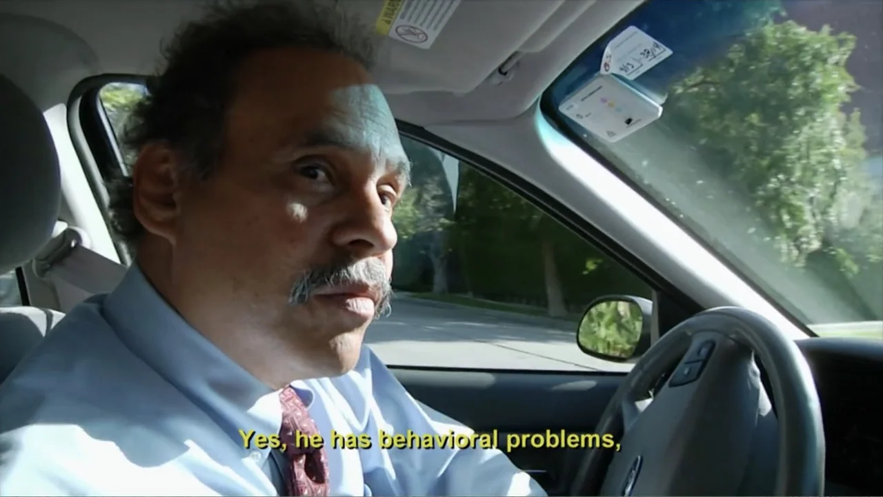A Dangerous Son (2018) movie still: closeup — A man sits in the driver seat of a moving car during the day. The man looks toward the passenger seat…