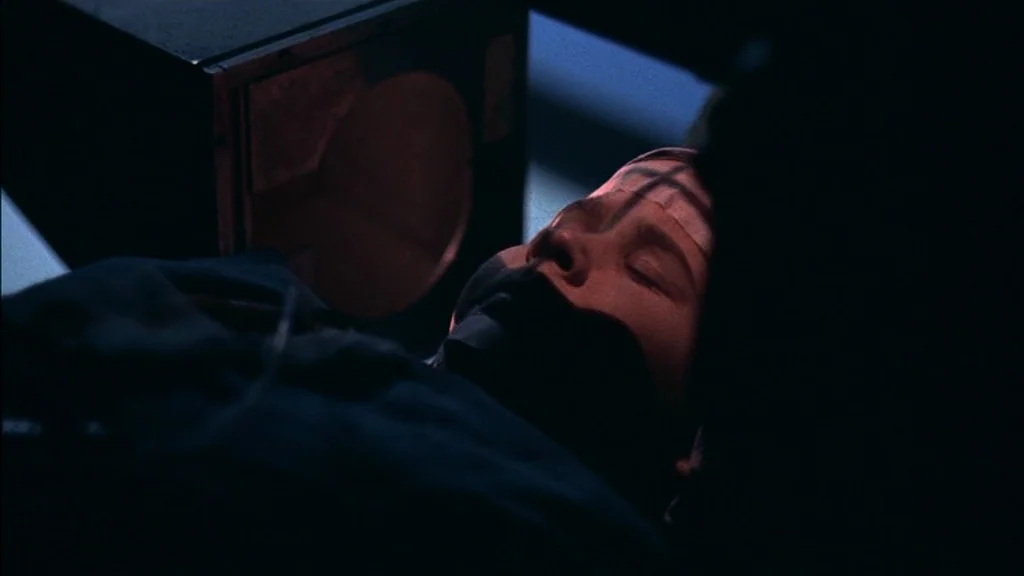 The Exorcist (1973) movie still: closeup — A dimly lit interior space contains a person lying on a flat surface. The subject has their mouth covered…