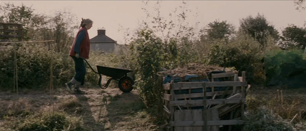 Another Year (2010) movie still: fullbody — A rural plot of land contains overgrown vegetation and a wooden compost bin structure. An elderly woman…
