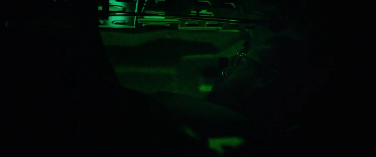 Uncut Gems (2019) movie still: closeup — A dark vehicle interior is lit only by a sharp green dashboard light source. A young man reclines in the…