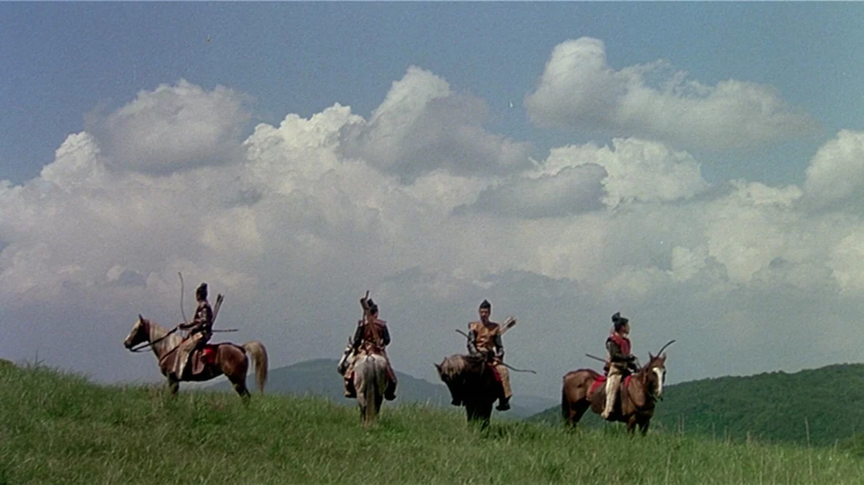 Ran (1985) movie still: wide — A grassy hill under a bright sky with large white clouds serves as the background. Four mounted archers in…