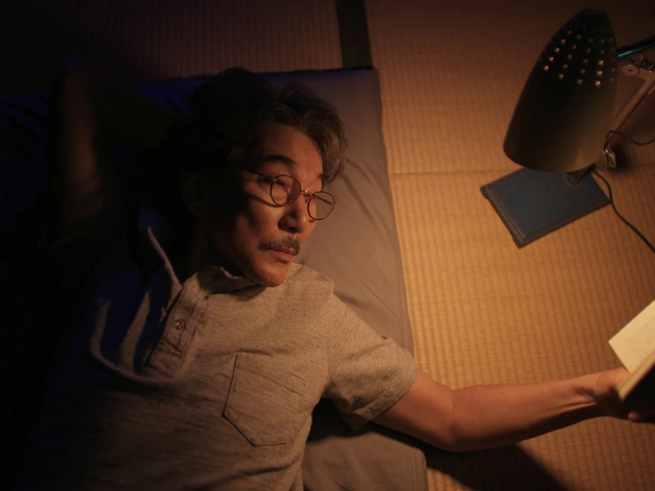 Perfect Days (2023) movie still: medium — A man lies on a traditional tatami floor inside a dimly lit room at night. He rests with one hand behind…