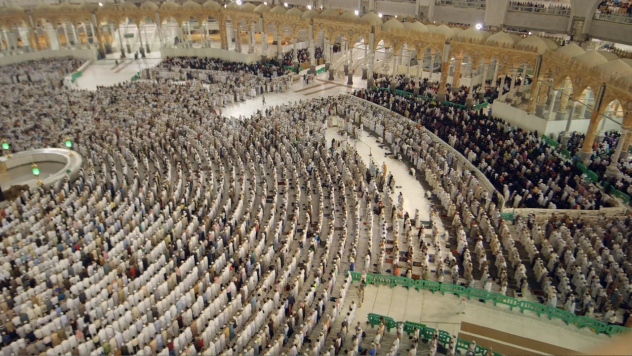 One Day in the Haram (2017) movie still: establishing — A vast outdoor courtyard with ornate golden arches contains thousands of people. The…