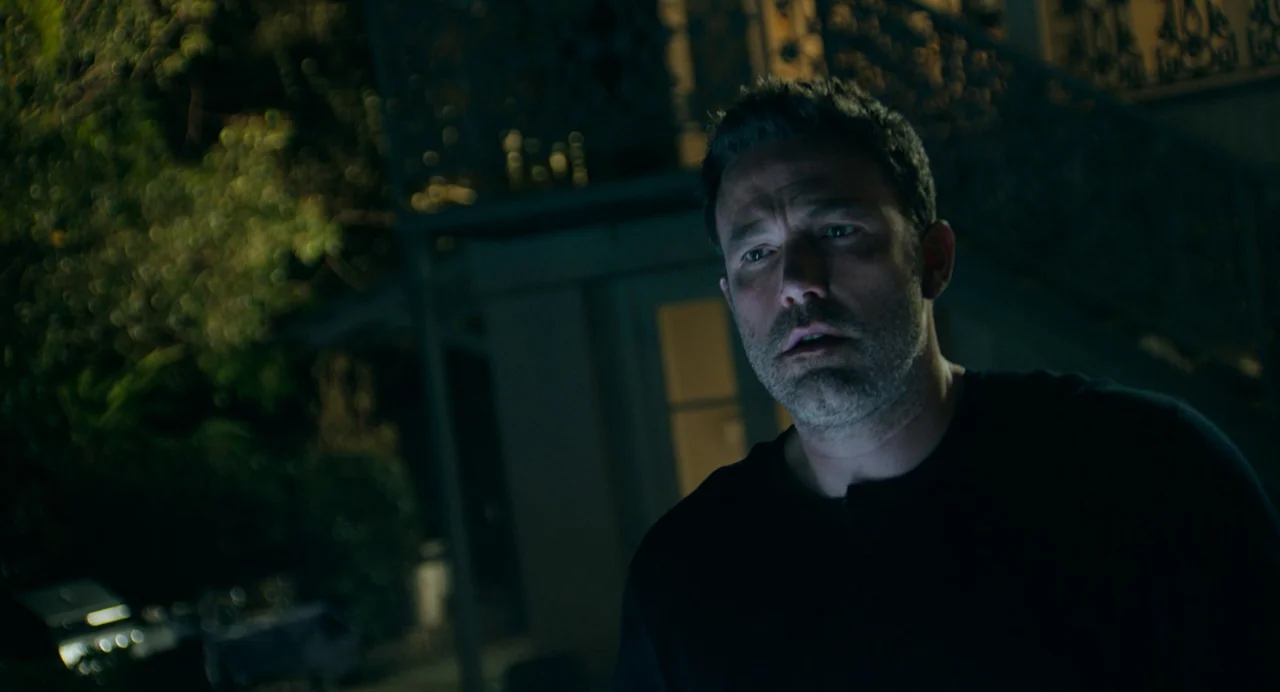 Deep Water (2022) movie still: medium — A dark, blurred backyard environment features an iron railing and foliage in the distance. A middle-aged man…