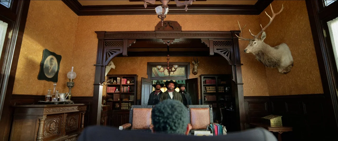 The Harder They Fall (2021) movie still: wide — A wood-paneled room features taxidermy on the walls and built-in bookshelves. Three men wearing hats…