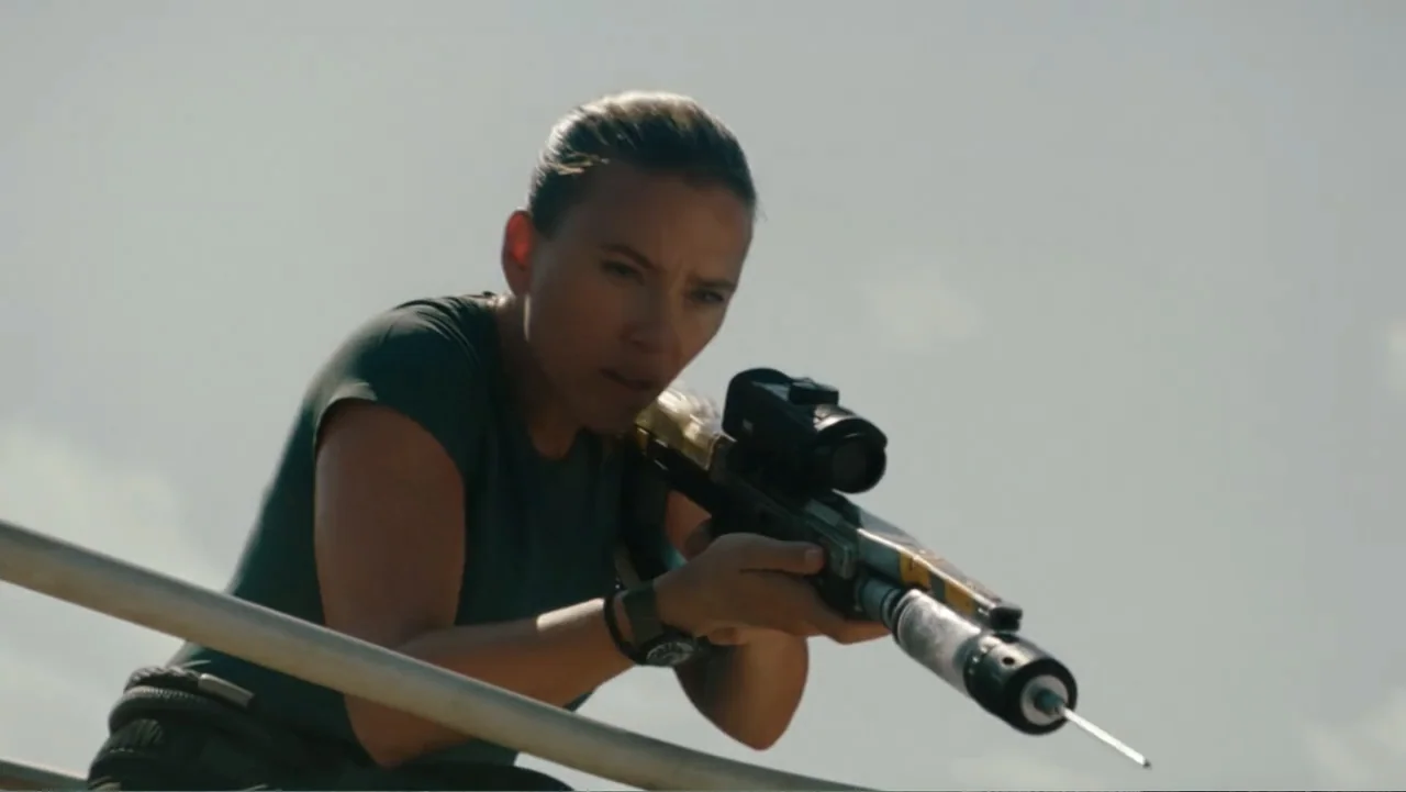 Jurassic World Rebirth (2025) movie still: medium — A woman stands on an outdoor metal railing platform against a bright, overcast sky. She holds…