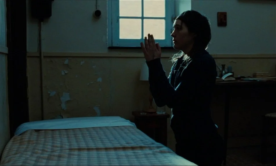 Hadewijch (2009) movie still: medium — A sparsely furnished room features a simple bed with a plaid blanket and a window. A woman kneels beside the…