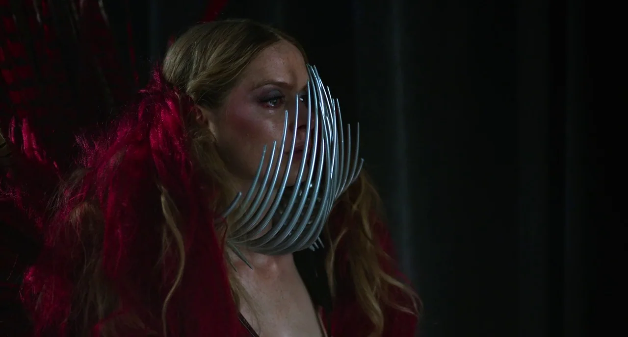 Rabid (2019) movie still: closeup — A dark space with a person wearing elaborate red textures and a metallic neck cage. The subject stands against a…