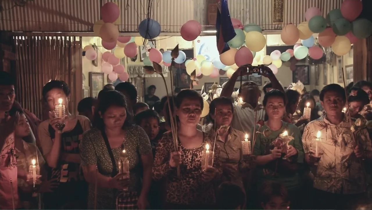 A Cambodian Spring (2018) movie still: medium — A crowded interior space is decorated with clusters of colorful hanging balloons. A diverse group of…