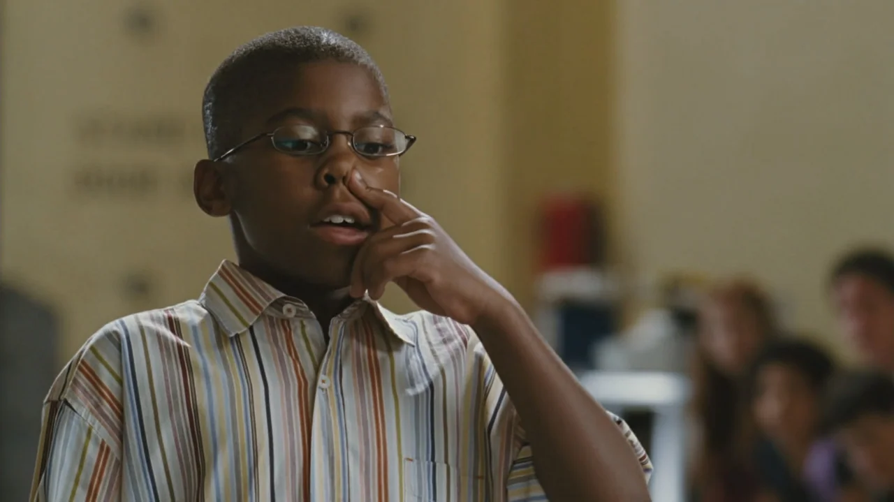 Zoom (2006) movie still: medium — A student stands in a brightly lit classroom with indistinct figures in the background. A young boy in a striped…