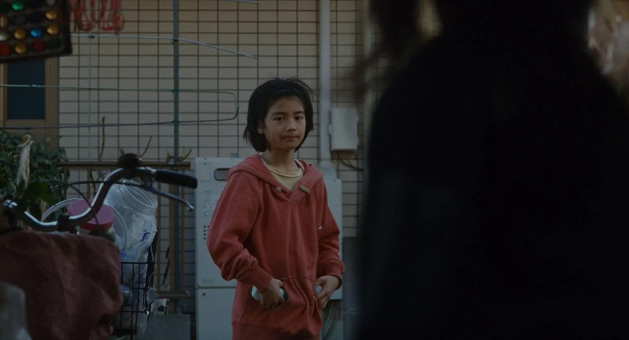 Shoplifters (2018) movie still: medium — A child stands outside a tiled residential building near a bicycle and yard clutter. The child looks toward…