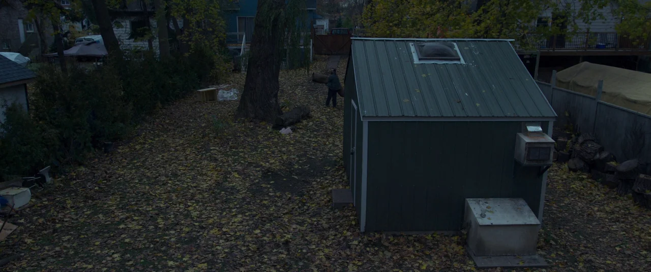 Room (2015) movie still: wide — A residential backyard covered in fallen autumn leaves with a green metal shed. A man drags a large log across the…