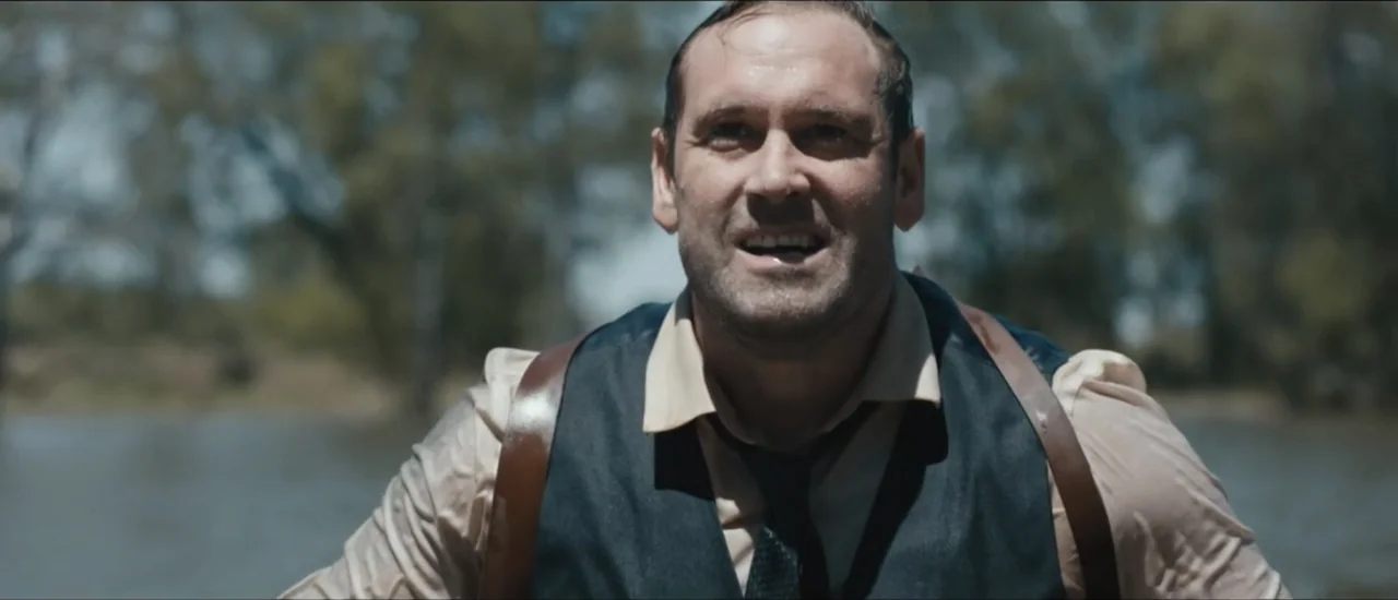 Fear Below (2025) movie still: medium — A man stands near a riverbank with trees visible in the soft-focus background. The man faces the camera…