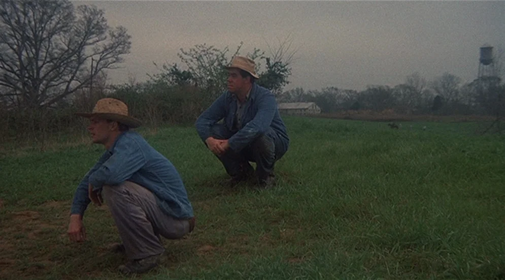 Thieves Like Us (1974) movie still: wide — An overcast day in a rural landscape with sparse grass and leafless trees. Two men wearing straw hats and…