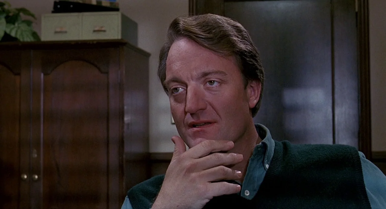 Child’s Play 2 (1990) movie still: closeup — A man sits in an office with dark wooden cabinets and a potted plant. He supports his chin with his…