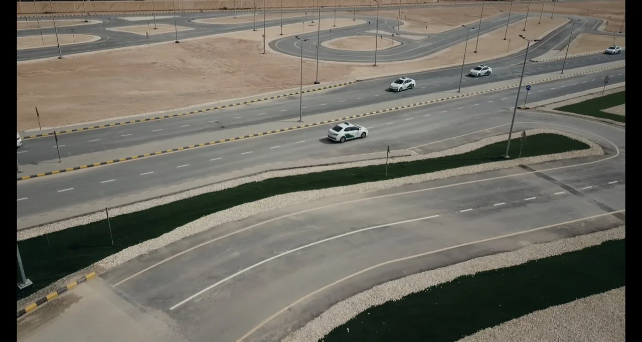 Saudi Women's Driving School (2019) movie still: wide — An extensive network of paved driving paths winds through a flat desert landscape. Multiple…