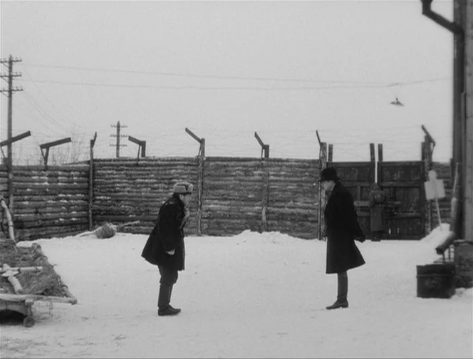 The Ascent (1977) movie still: wide — A snowy yard enclosed by a tall log wall topped with barbed wire. Two men stand facing each other in the…