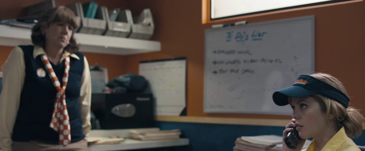 Compliance (2012) movie still: medium — An office inside a fast food restaurant features beige walls and a white board. Two women stand in the…
