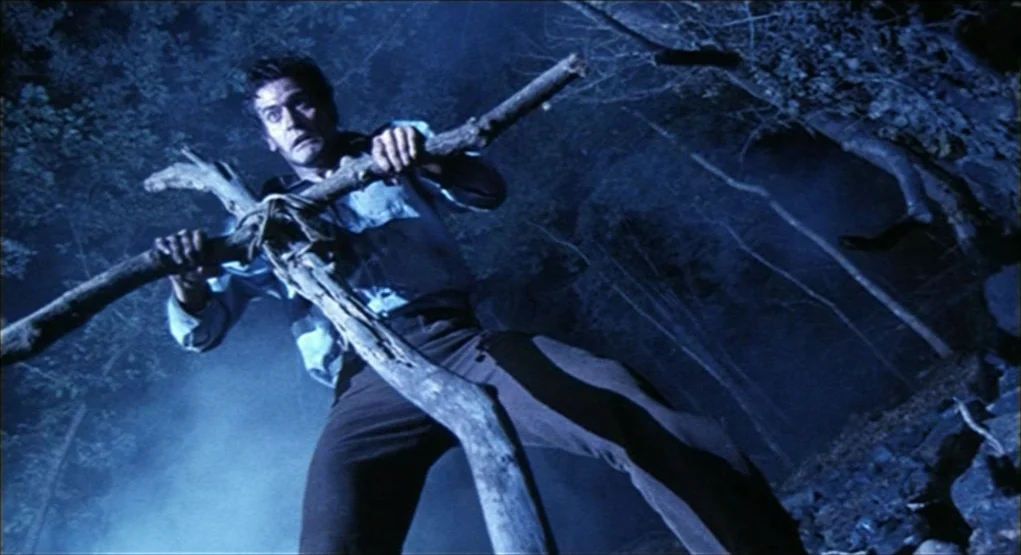 Evil Dead II (1987) movie still: fullbody — A dark night setting within a dense woods with thick foliage and low light. A man holds a large wooden…