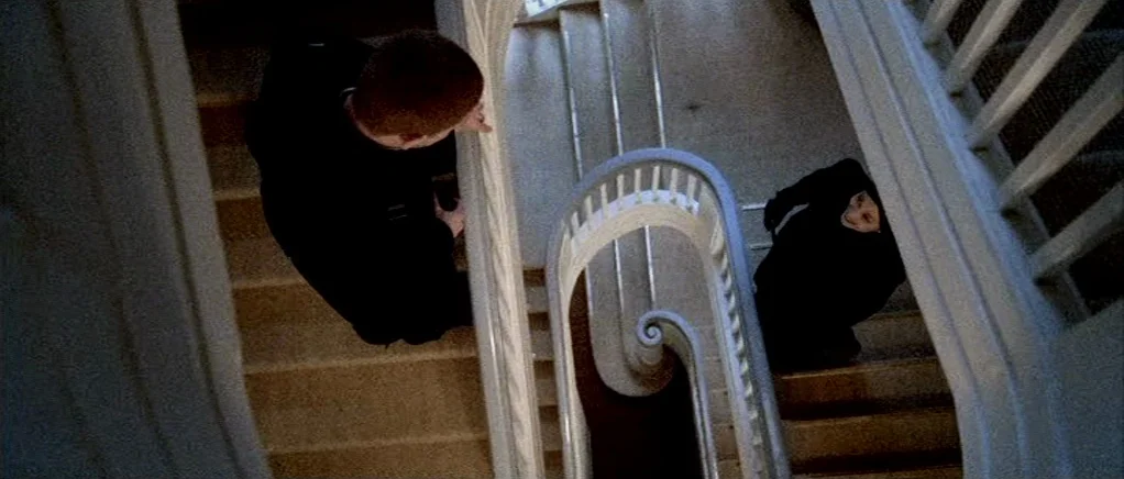 Reconstruction (2003) movie still: wide — An interior spiral staircase with white balusters curves down through multiple levels. Two figures dressed…