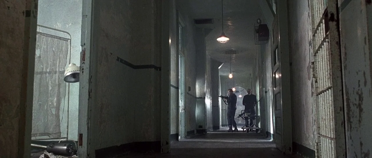 The Rock (1996) movie still: wide — A long, dimly lit hospital corridor features peeling green paint and metal bar gates. Two men in dark clothing…