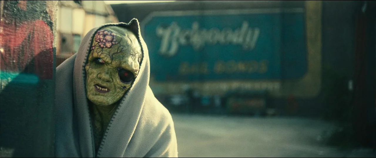 The Toxic Avenger (2023) movie still: medium — An outdoor urban space features a large faded sign for a bail bonds business. A green skinned…
