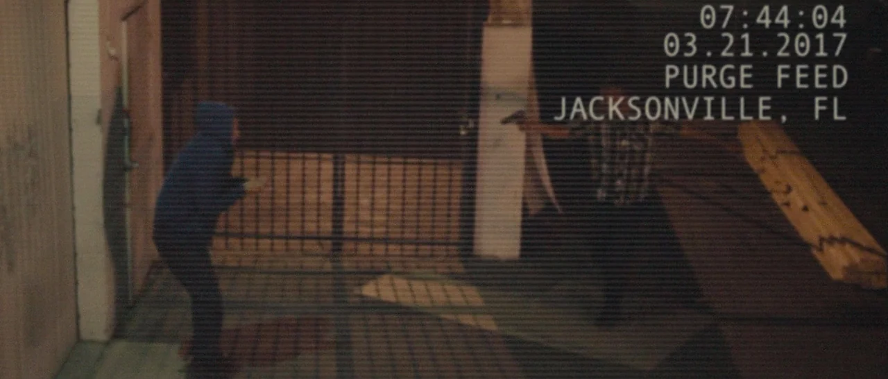The Purge (2013) movie still: wide — A dimly lit concrete structure shows surveillance overlay text in the top right corner. Two figures stand in a…