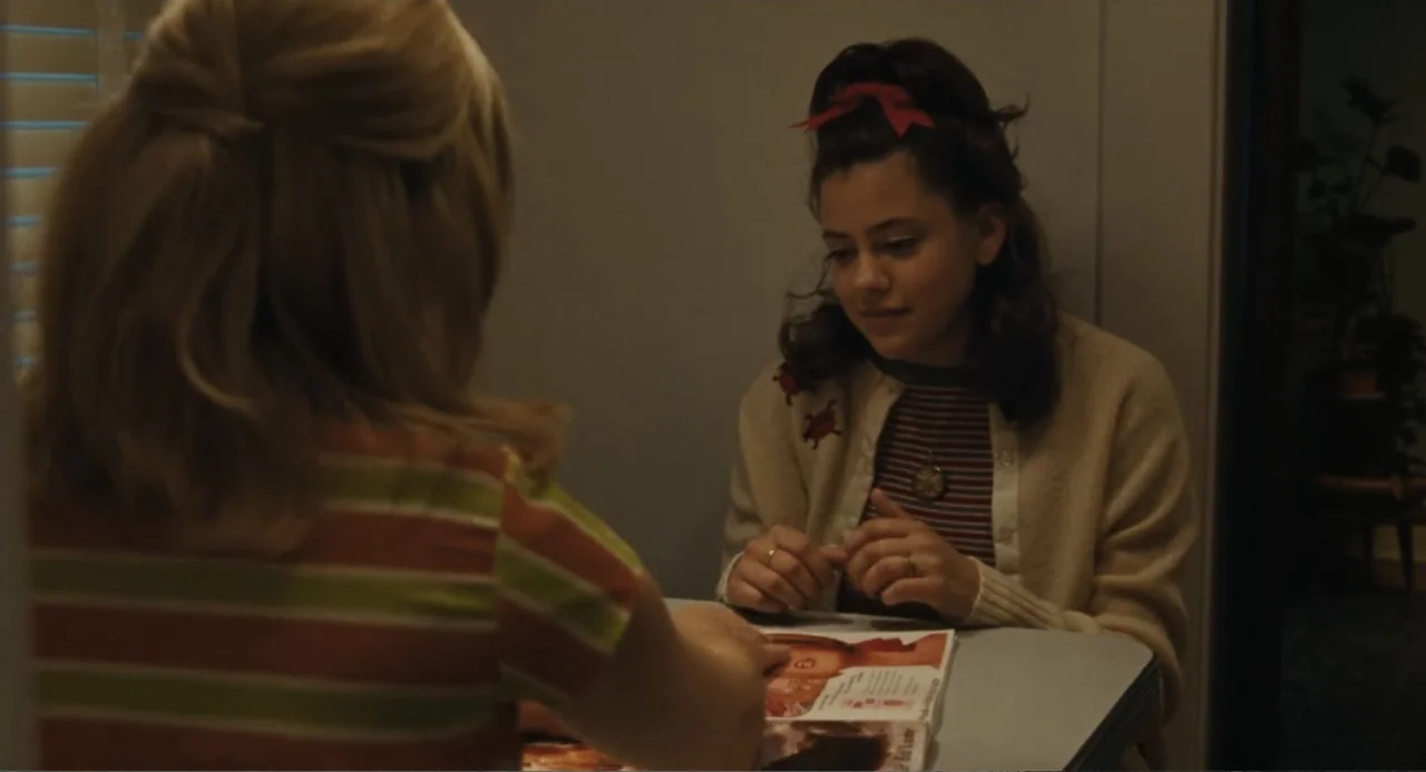 Call Jane (2022) movie still: medium — Two girls sit across from each other at a diner table with patterned walls. One girl wears a cream cardigan…