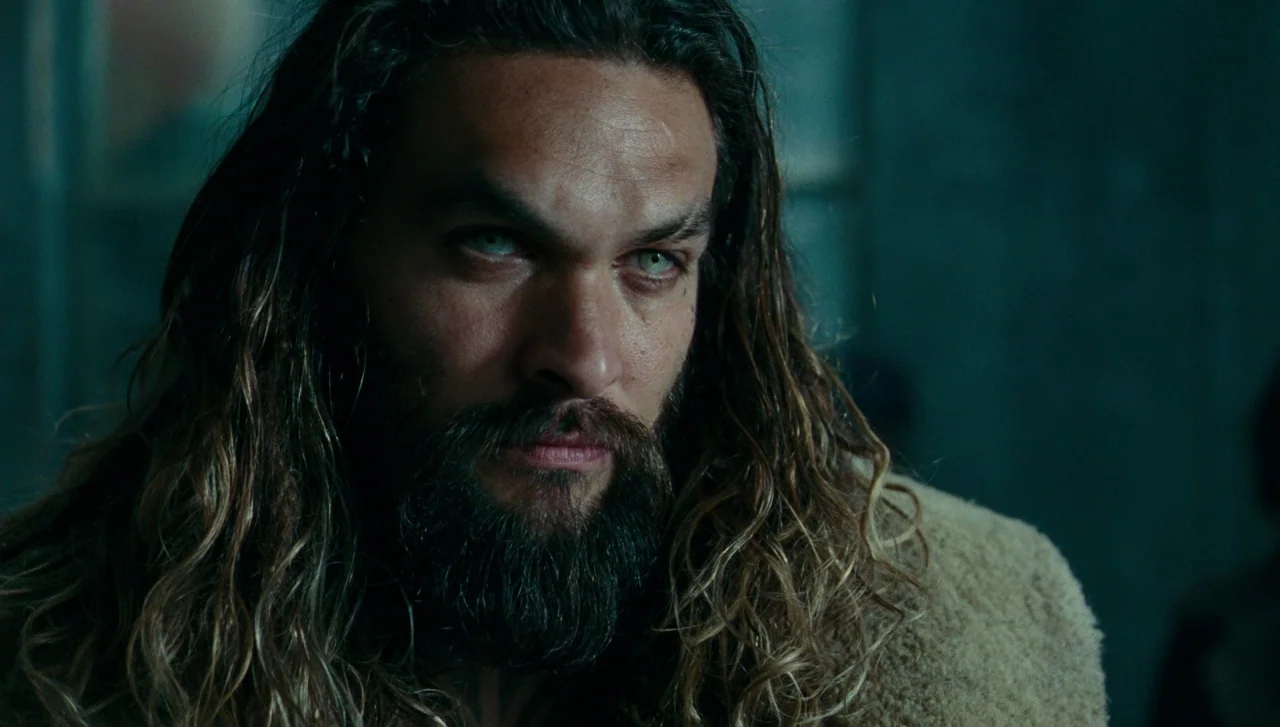 Justice League (2017) movie still: closeup — A dimly lit interior space with cold teal tones and deep background shadows. A man with long hair and a…