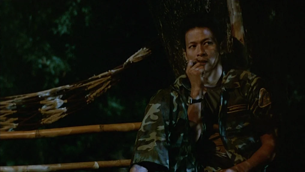 Tropical Malady (2004) movie still: medium — A dark jungle environment at night with a tree and bamboo railing visible. A man in camouflage gear…